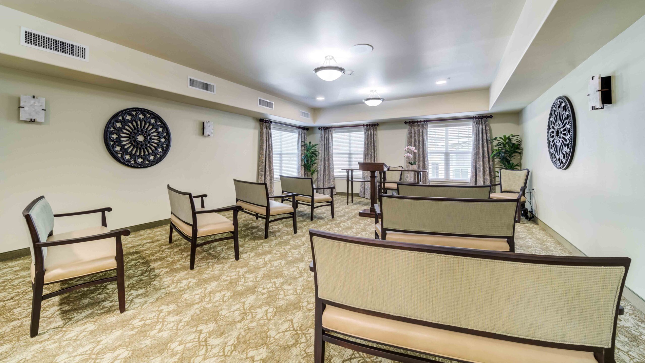 A small, well-lit room with several cushioned chairs arranged in a semi-circle facing a podium. There are plants, curtains on large windows, and decorative wall art. The space appears clean and organized.
