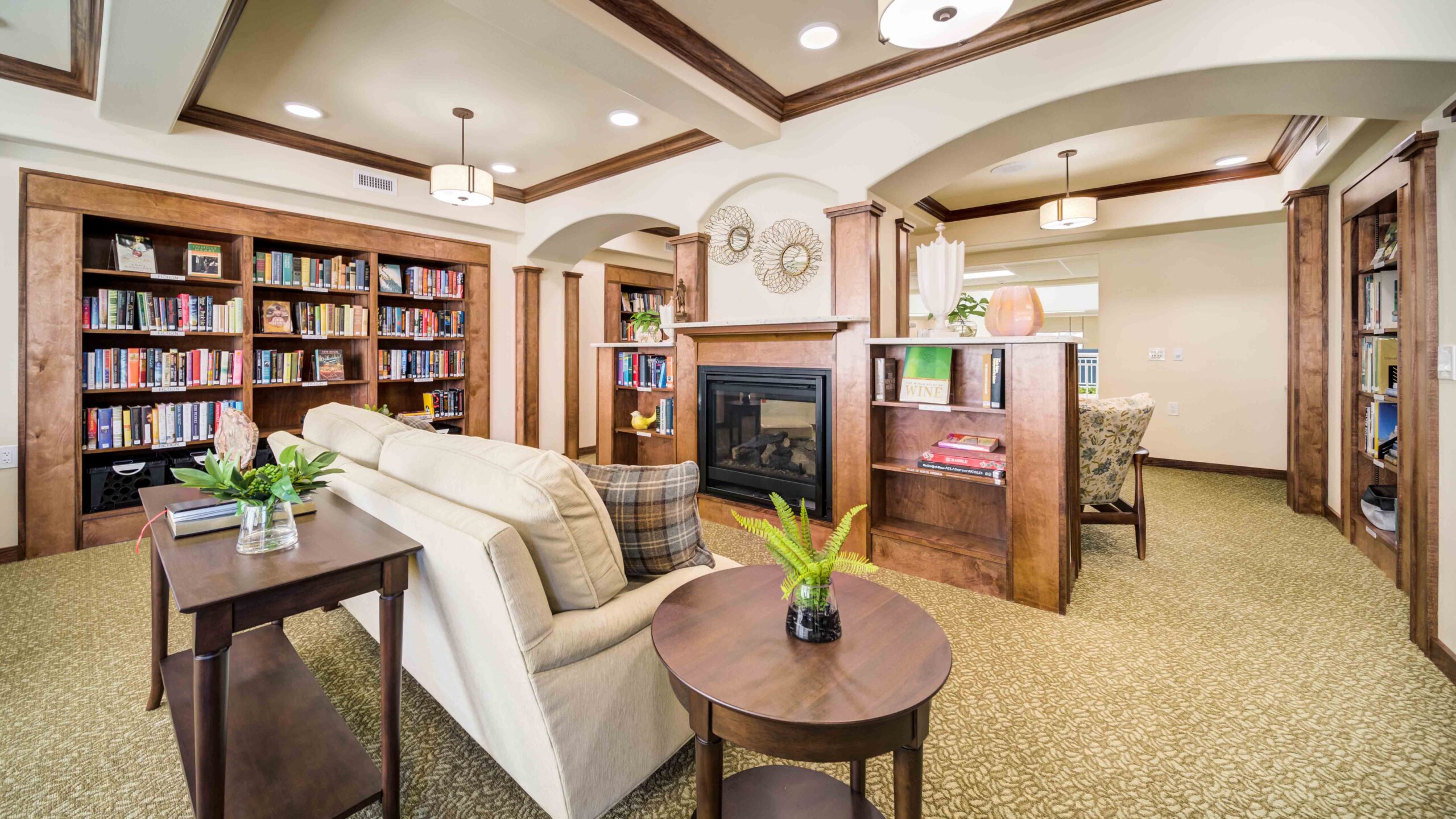 A cozy library room with bookshelves, couches, armchairs, and a central fireplace. Decorative plants and ornaments are on tables and shelves, and arched doorways open to other areas. The space is warmly lit and inviting.