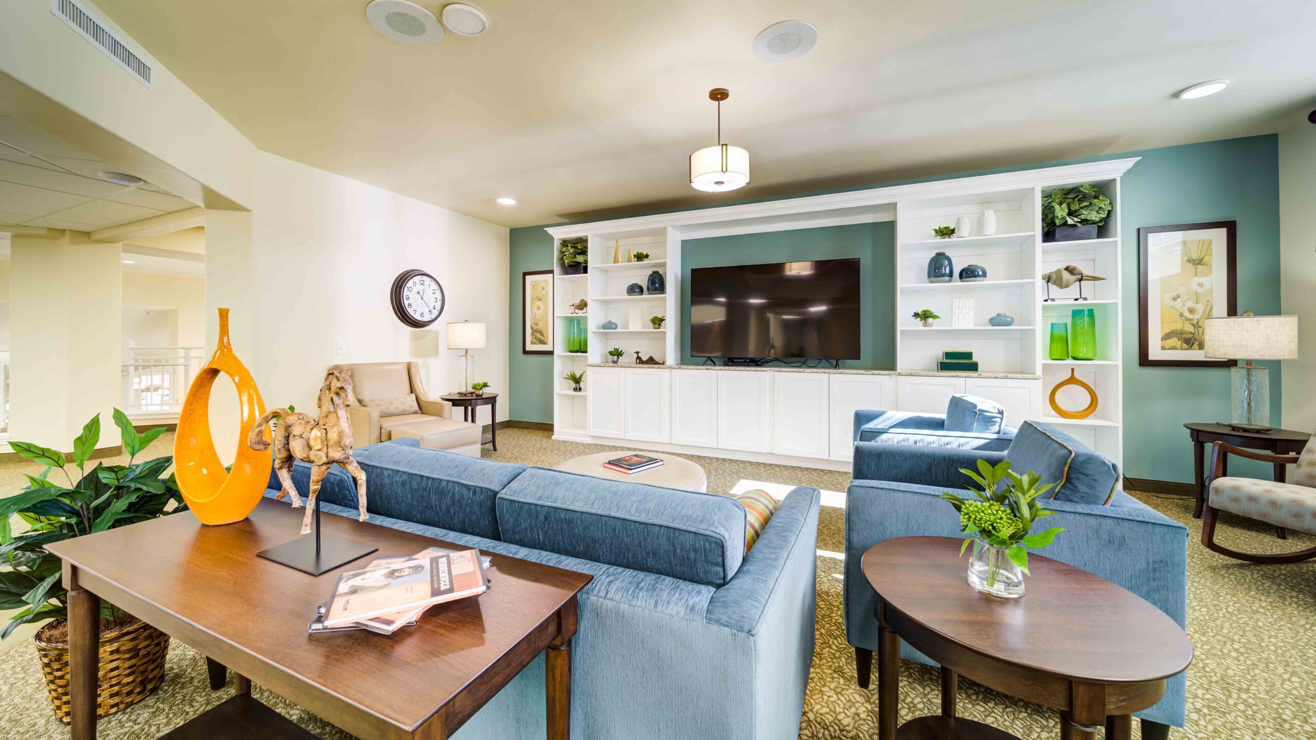 A modern, cozy living room with blue sofas, a large TV on a white built-in shelving unit, decorative plants, books, and art pieces. Soft lighting and a mix of contemporary and classic decor create a welcoming space.