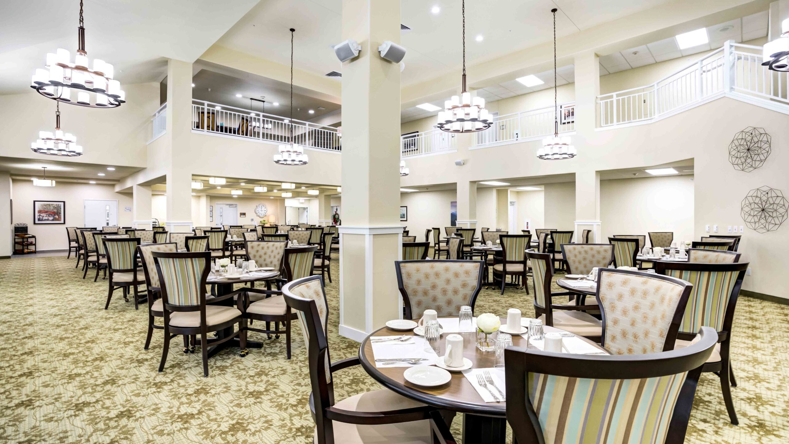 A spacious, well-lit dining room with round and square tables set for meals, cushioned chairs, chandeliers, patterned carpet, and a balcony overlooking the area. The setting appears elegant and inviting.