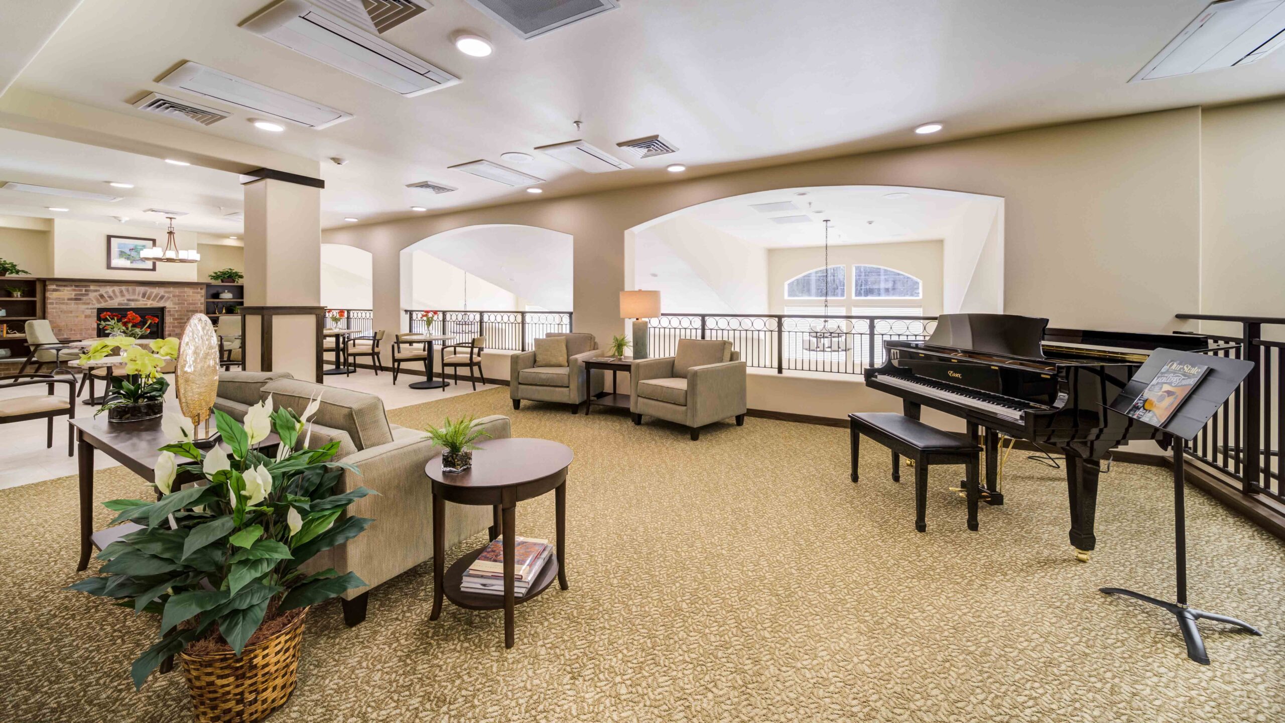 Bright, spacious lounge with beige carpeting, comfortable armchairs, potted plants, a grand piano with a music stand, and large arched windows letting in natural light. Dining area visible in the background.