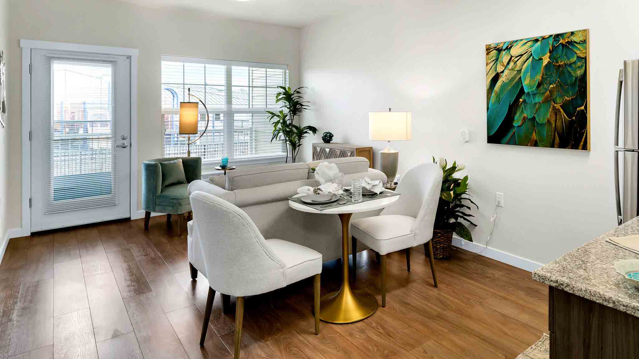 Modern living and dining area with wooden floors, a round table set for four, light-colored chairs, a sofa, large windows, indoor plants, a floor lamp, and a colorful feather artwork on the wall.