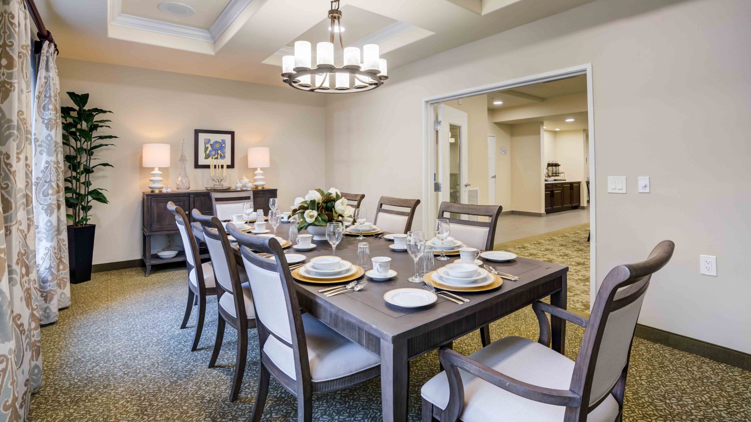 A dining room with a dark wooden table set for eight, elegant upholstered chairs, a chandelier above, sideboard with lamps, framed art, potted plant, and open double doors to another room.