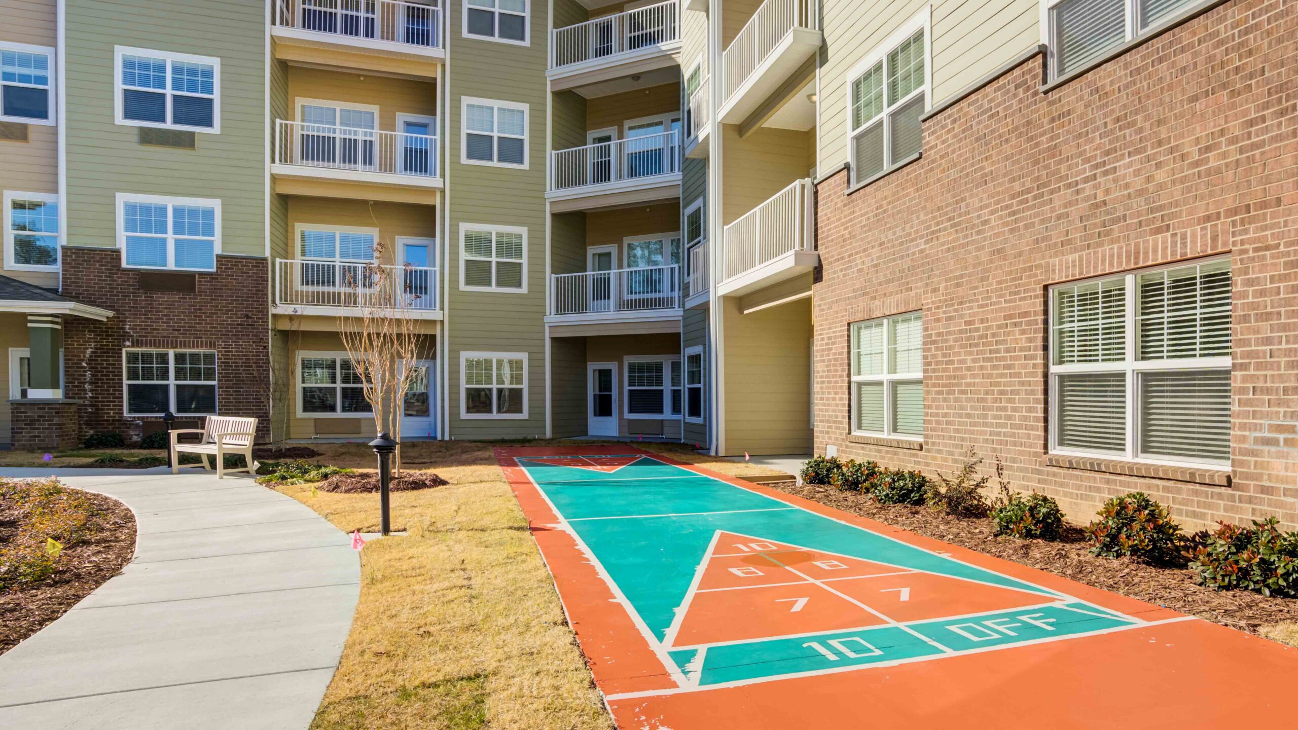 A shuffleboard court is painted on the ground in the courtyard of an apartment complex, surrounded by landscaped areas, a walkway, benches, and several apartment balconies.