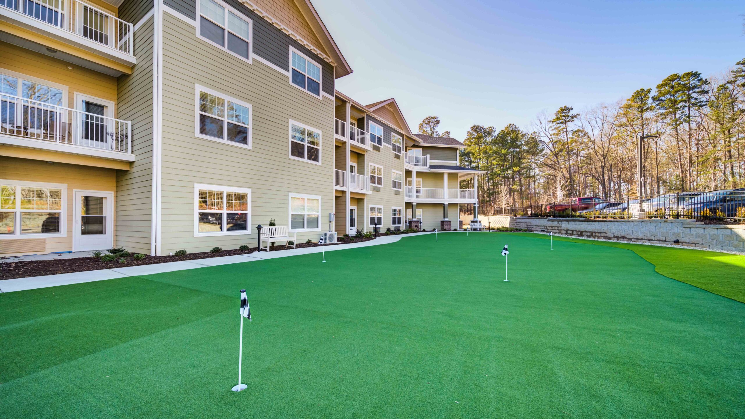 A modern apartment building with beige siding overlooks a well-maintained artificial putting green with several golf holes and flags, bordered by a sidewalk and surrounded by trees.