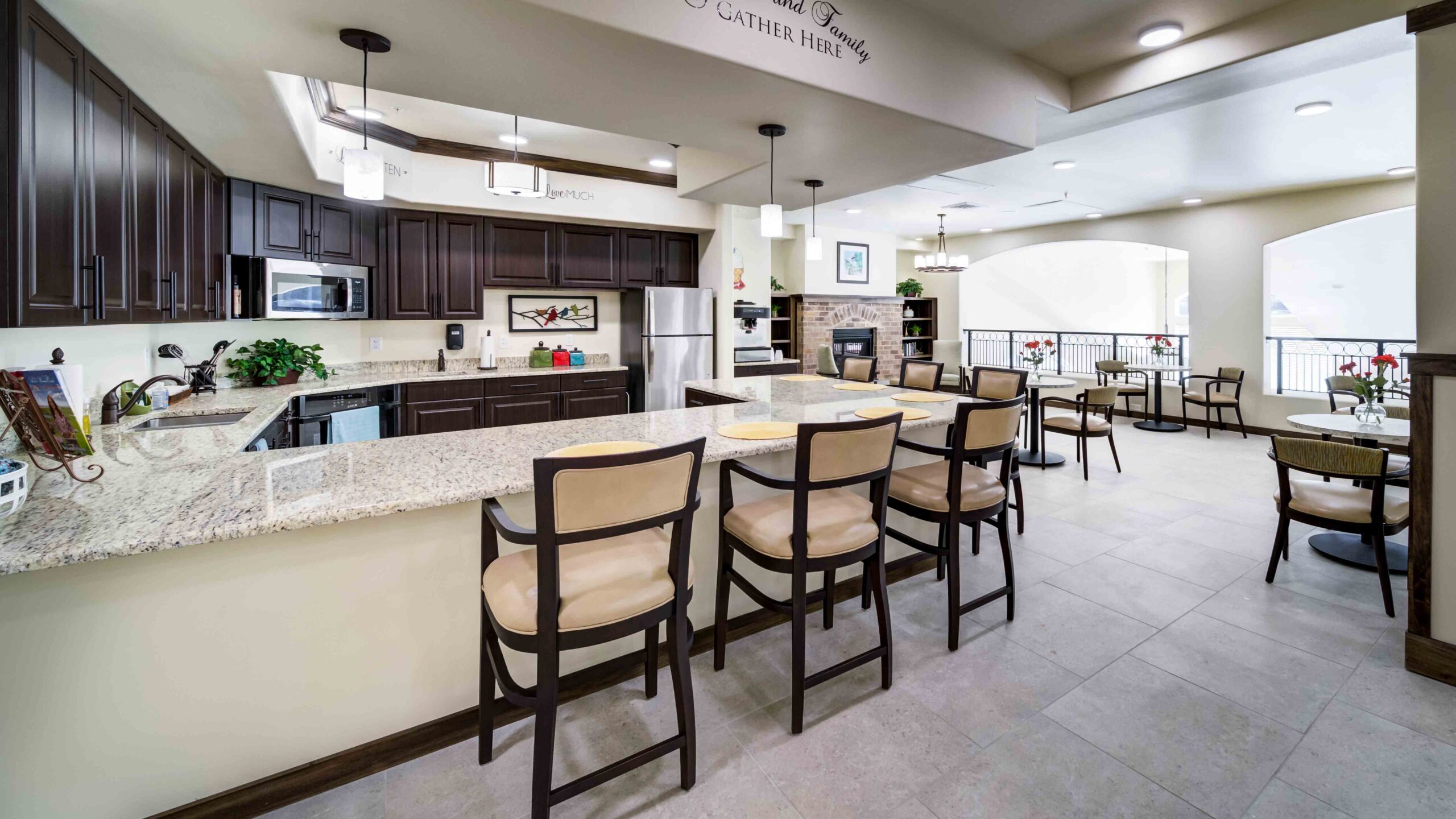 A spacious, modern kitchen with dark cabinets, stainless steel appliances, granite countertops, and a breakfast bar with six chairs. Nearby, several round dining tables and chairs are arranged in a bright, open area.