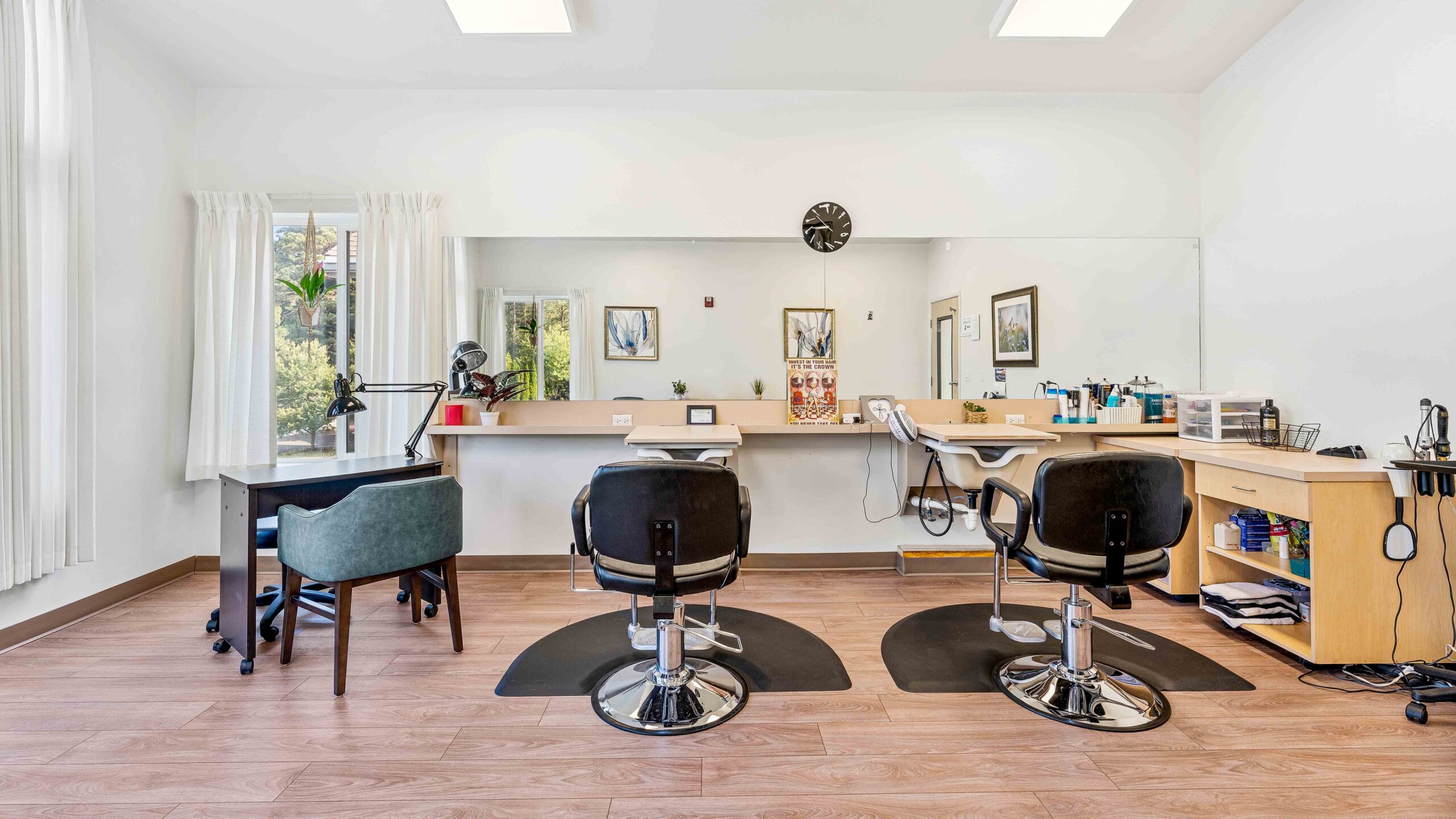 A bright, modern salon with two black styling chairs facing a large mirror, workstations on either side, a desk with a green chair, wooden floors, and white walls with minimal decor.