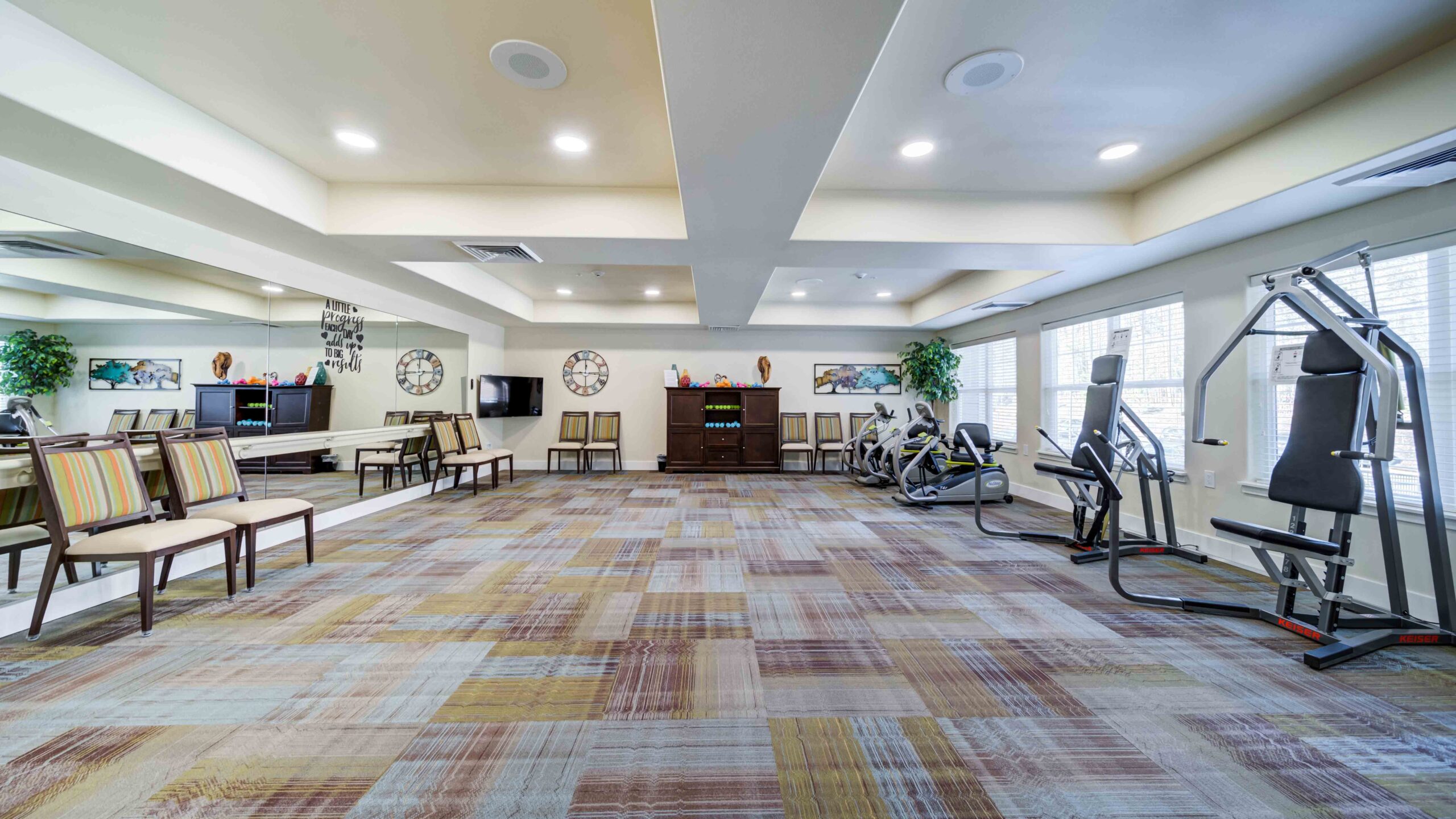 Spacious fitness room with patterned carpet, exercise machines on the right, chairs lined up on the left, large windows letting in natural light, and neutral-colored walls with ceiling lights.