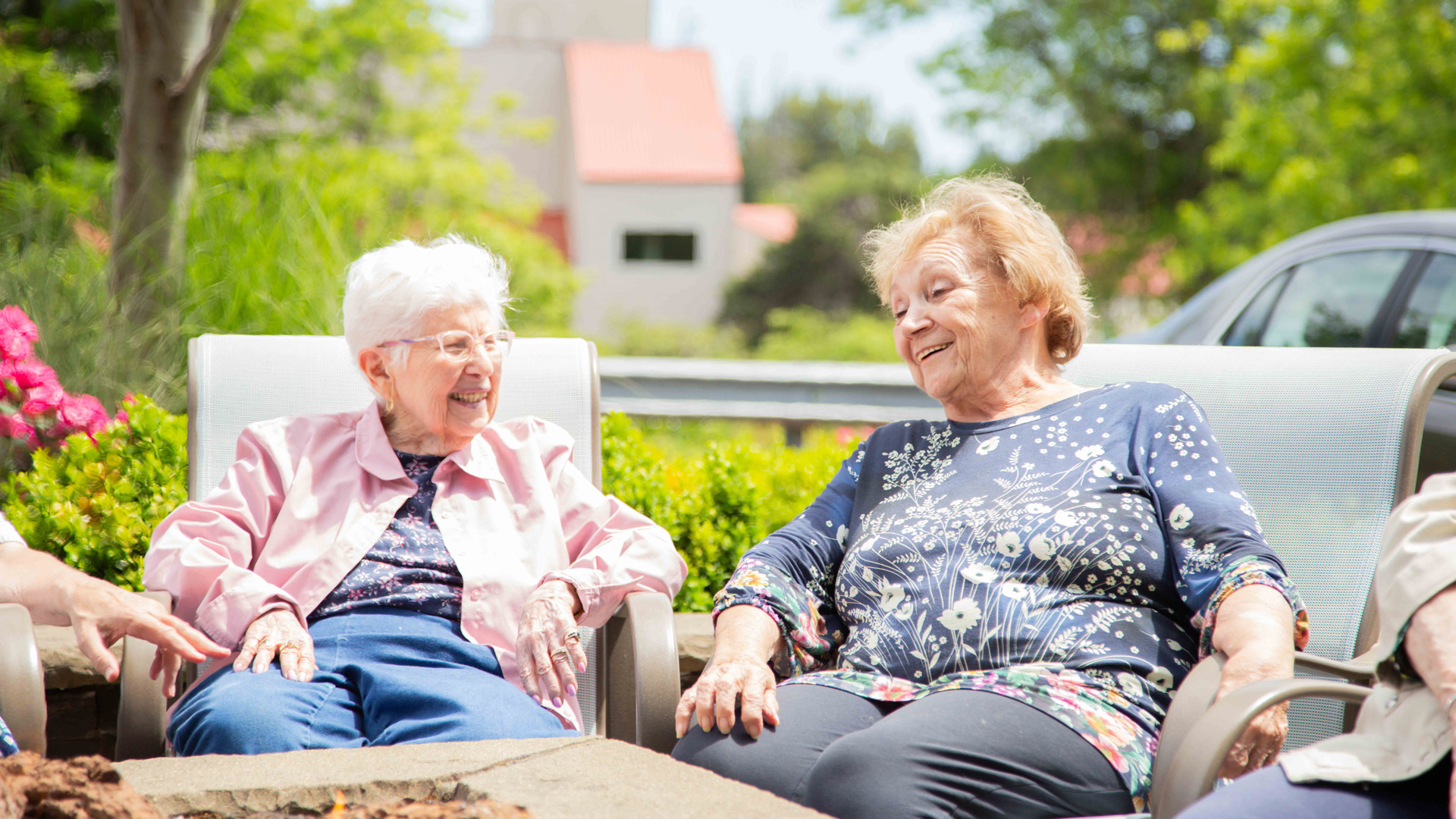 Two elderly women sit outdoors on patio chairs, smiling and laughing together on a sunny day, surrounded by greenery and flowers.