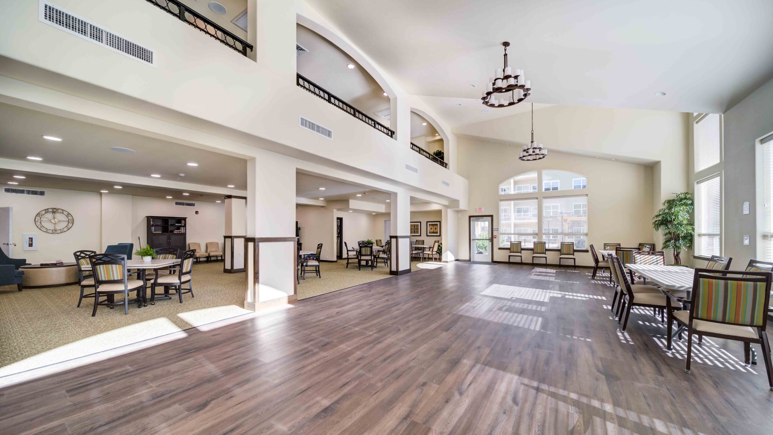 A spacious, well-lit dining and common area with high ceilings, large windows, wooden floors, tables, and chairs arranged neatly. The room features modern décor and double-height space with a lofted overlook.