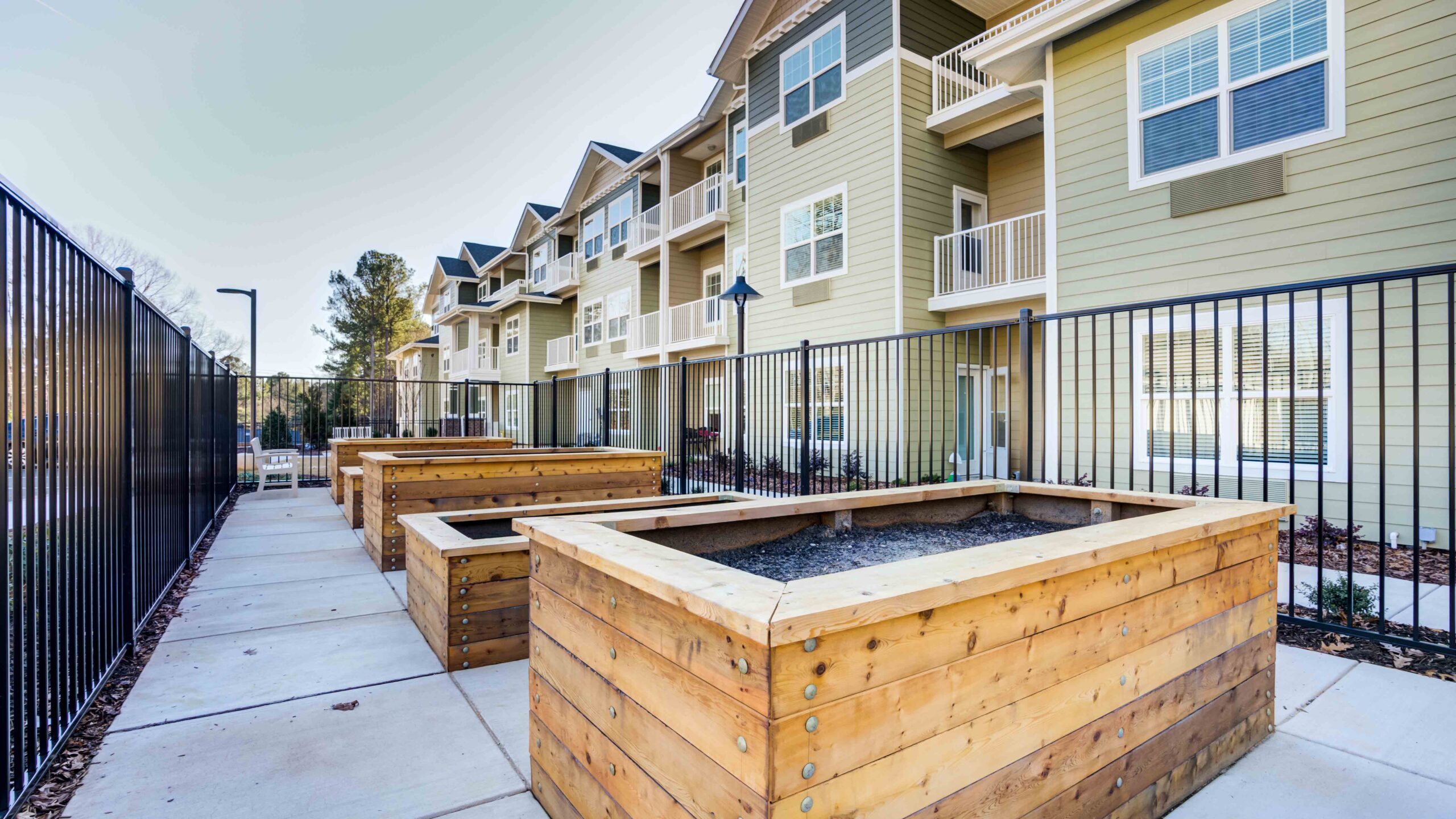 Raised wooden garden beds filled with soil line a paved walkway beside a fence, in front of a modern apartment building with balconies and large windows.