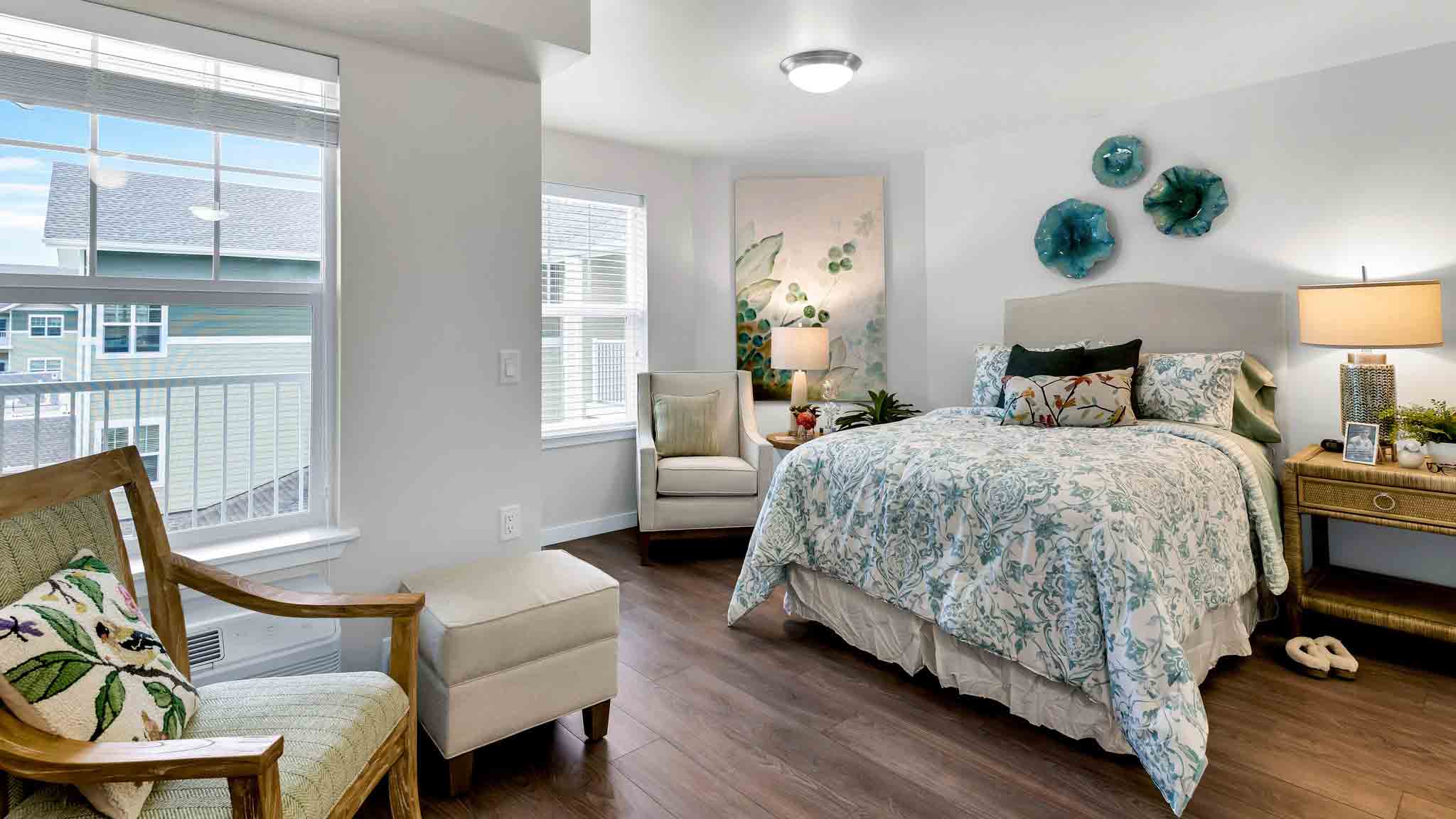 A bright, cozy bedroom with a floral-patterned bed, a beige armchair, wooden chair, nightstand with a lamp, and green wall decor. Large windows let in natural light, and the room has wood flooring.