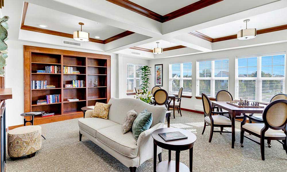 A bright, cozy communal room featuring a beige sofa with cushions, round wooden side tables, and a bookshelf filled with books against the wall. Several wooden tables and chairs are scattered around the room, with large windows allowing natural light to flood in.