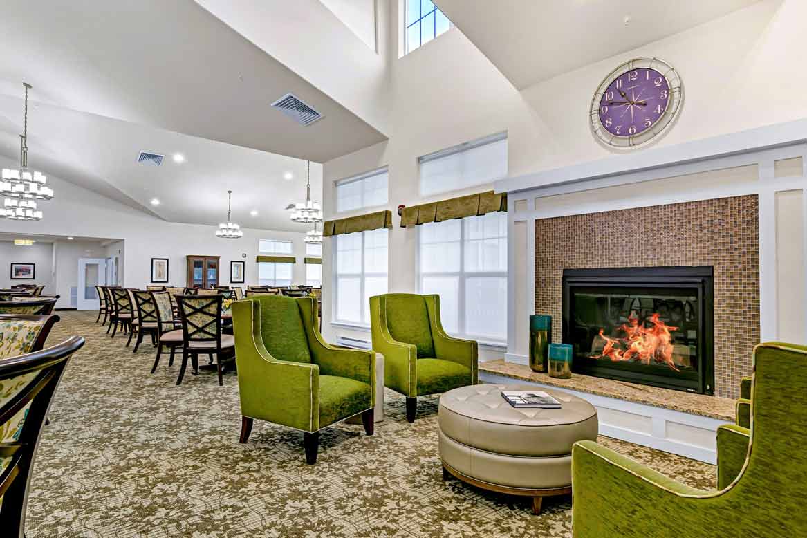 A well-lit lounge area with green armchairs and a circular ottoman in front of a fireplace. A large, purple clock hangs above the fireplace. The room has many chairs and tables, patterned carpet, and large windows with white shades.