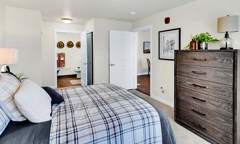 A cozy bedroom with a bed covered in plaid bedding, a wooden dresser with plants and a lamp, and a nightstand with a lit lamp. The room has beige walls, a soft carpet, and an open door leading to a bathroom with towels and a wall art display.