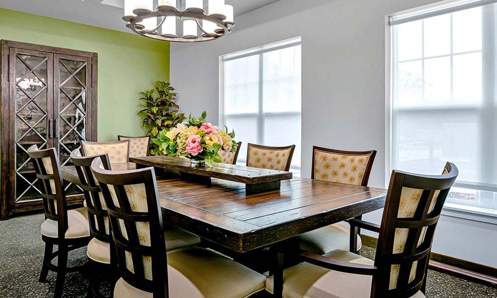 A dining room features a large wooden table with eight cushioned chairs. A colorful flower arrangement adorns the table. The room has a chandelier, a green wall with wooden cabinet, and windows with white shades that let in natural light.