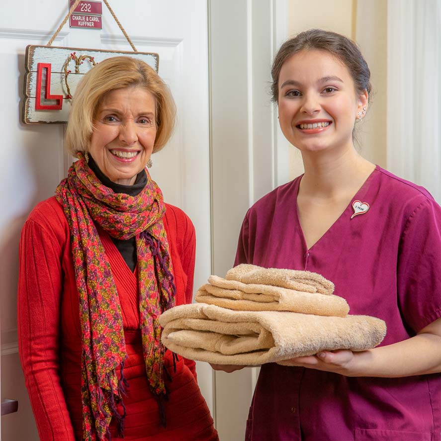 An elderly woman wearing a red top and colorful scarf stands next to a young woman in a maroon uniform holding folded towels. They are smiling in front of a door decorated with a sign that reads 