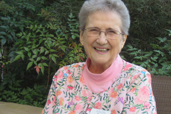 An elderly woman with short gray hair and glasses is smiling while sitting outdoors. She is wearing a pink floral-patterned top and a pink turtleneck. The background features lush green foliage.
