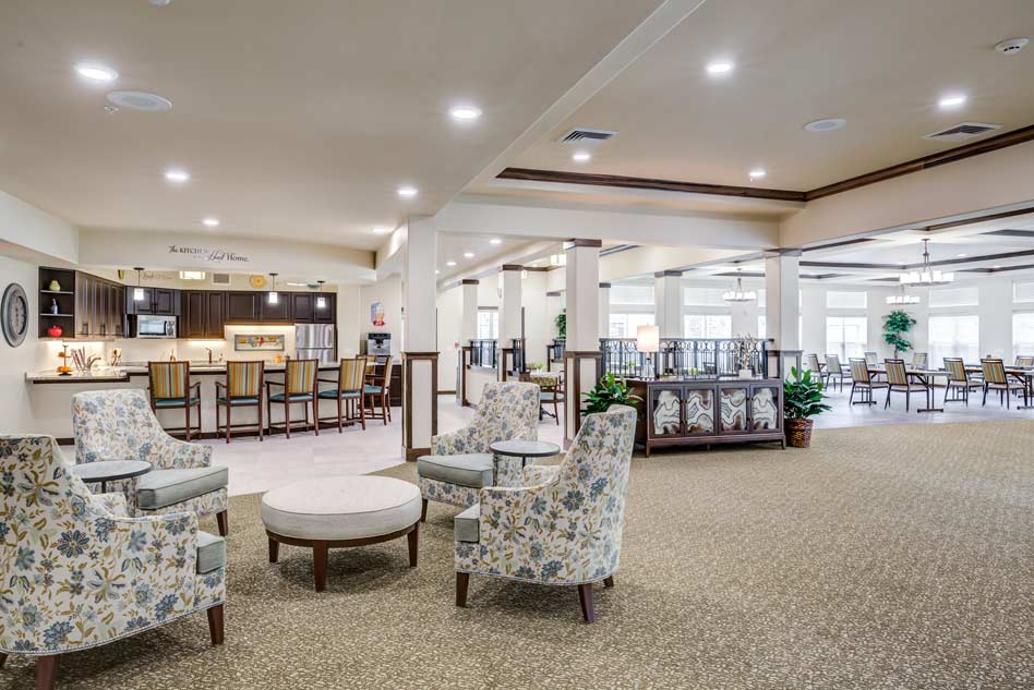 A spacious communal area features a seating arrangement with floral-patterned armchairs and a circular coffee table. In the background, there is a countertop bar with stools and a large dining area with tables and chairs. The space is well-lit and welcoming.