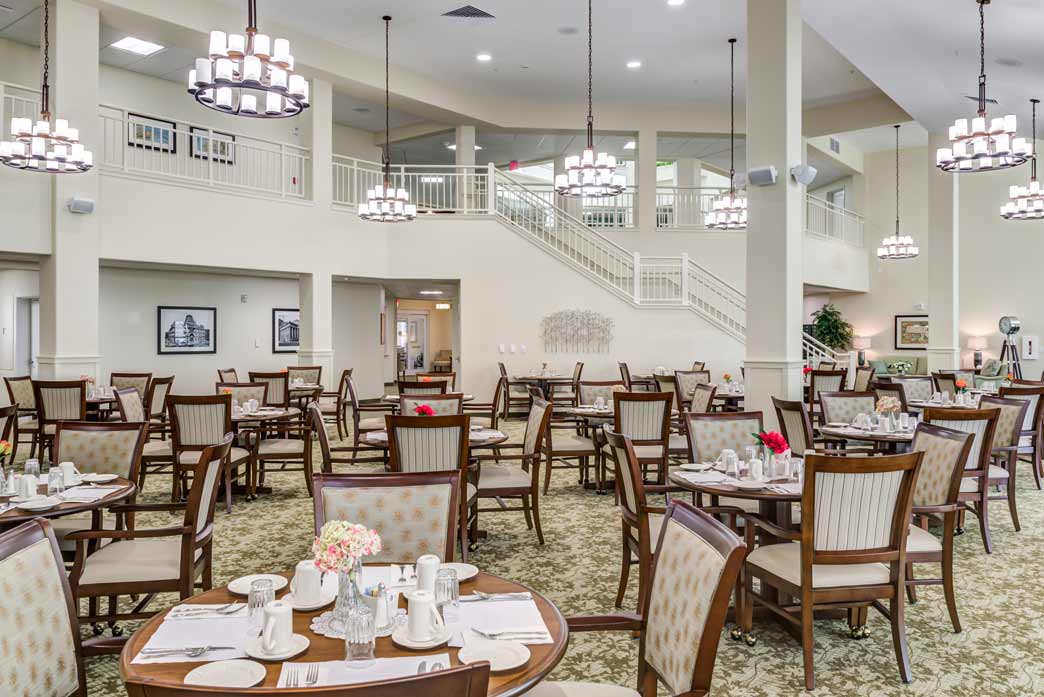 A spacious dining room with high ceilings and large chandeliers, featuring several neatly arranged tables and chairs. Each table is set with plates, cups, and napkins. A staircase leads to an upper level, and artwork and plants decorate the room.