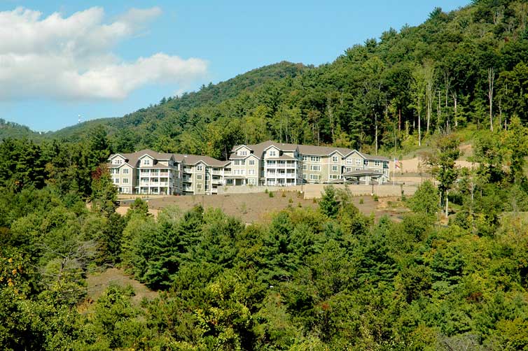 Large multi-story residential buildings with numerous windows sit atop a forested hill under a clear blue sky. Dense greenery from trees covers the hillside, providing a lush backdrop. White clouds are scattered in the sky, adding contrast to the landscape.