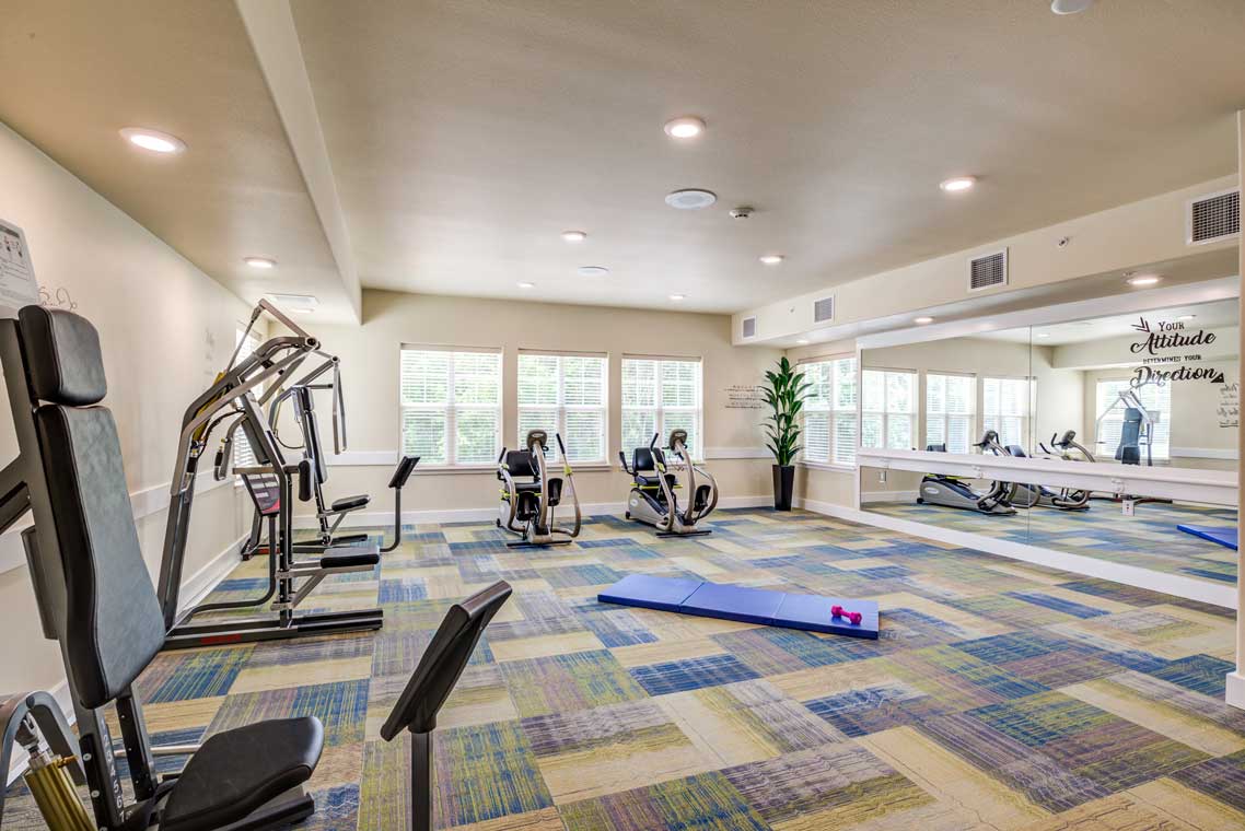 A brightly lit exercise room with various fitness equipment including weight machines, stationary bikes, and a yoga mat on the floor. A large mirror runs along one wall, with motivational quotes on it. The floor has a colorful geometric carpet pattern.