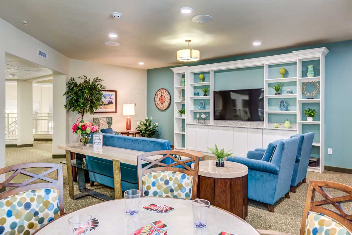 A cozy, modern living room with a blue and white color scheme. It features a large wall-mounted TV, blue sofas, a coffee table, built-in shelves with decorative items, a clock, and indoor plants. In the foreground, there is a dining table with colorful polka dot cushions.