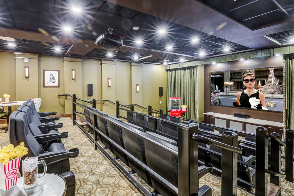 A cozy home theater room with recliner seating, a projector, and a large screen displaying a movie scene. The room has soft lighting, a popcorn machine, and framed pictures on the walls. Some seats have popcorn containers nearby.