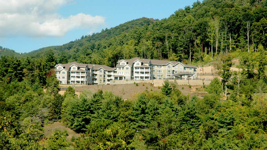 A large, gray building complex with multiple sections stands on a hillside surrounded by dense green trees and foliage. In the background, a forested mountain extends under a partly cloudy sky.