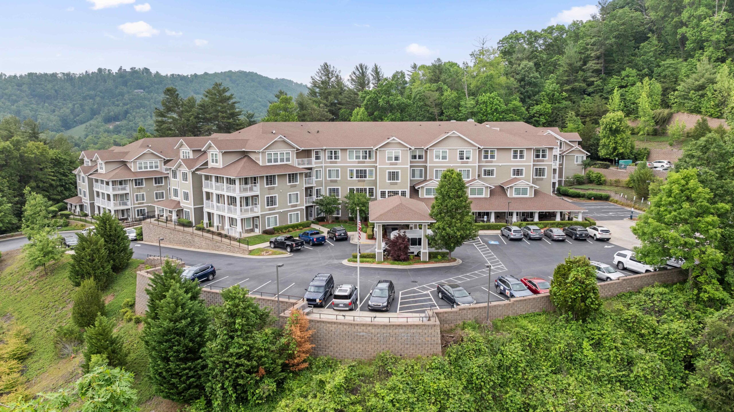 A large, multi-story residential building with beige siding and white trim sits on a hillside surrounded by lush green trees, with several cars parked in the lot in front and forested mountains in the background.