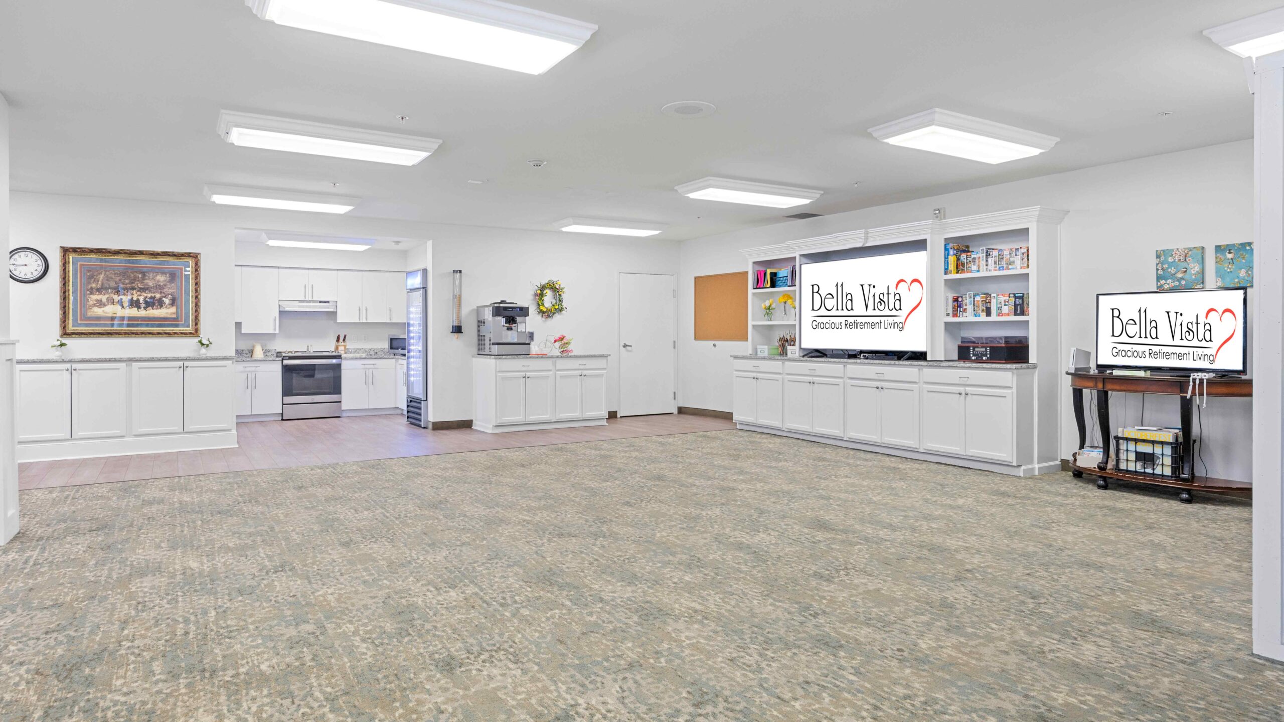 A spacious, well-lit communal area with carpeted floors, white cabinets, a kitchen, and two TVs displaying "Bella Vista". The room is decorated with art, books, and has a clean, organized appearance.