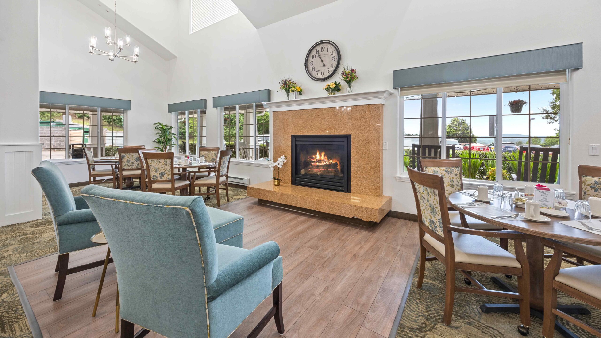A bright, elegant dining room with several tables and chairs, a fireplace with a clock above it, large windows letting in natural light, and decorative flowers on the mantel.