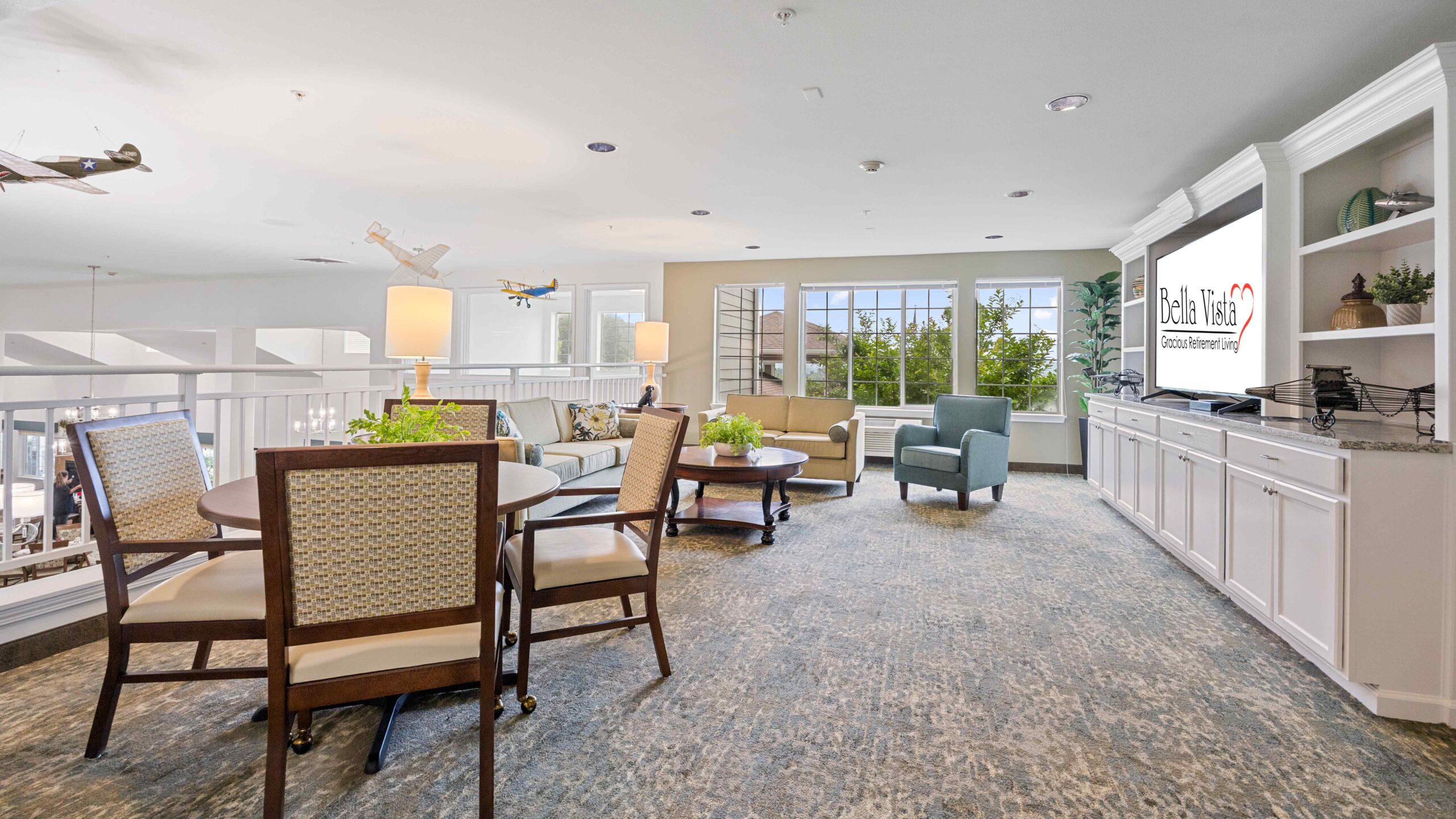 A bright, spacious lounge area with a round table and chairs, couches, armchairs, large windows, built-in shelves, and a TV displaying “Bella Tosa.” Model airplanes hang from the ceiling.