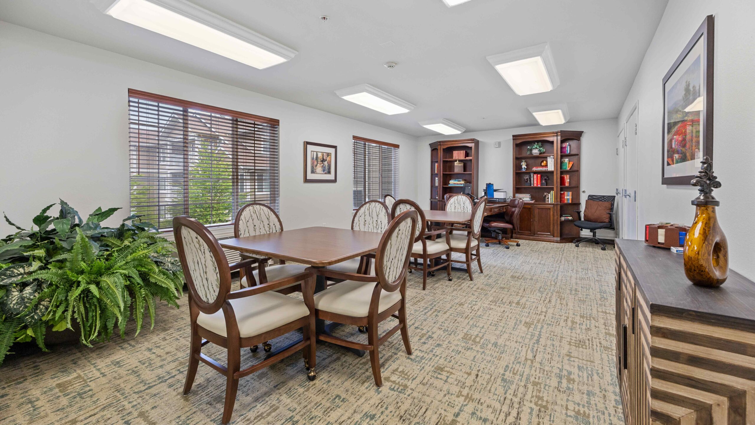 A well-lit office with two wooden tables and eight chairs, a large potted plant, bookshelves, framed art, and a desk with a computer in the background. The room has beige carpet and two windows with blinds.