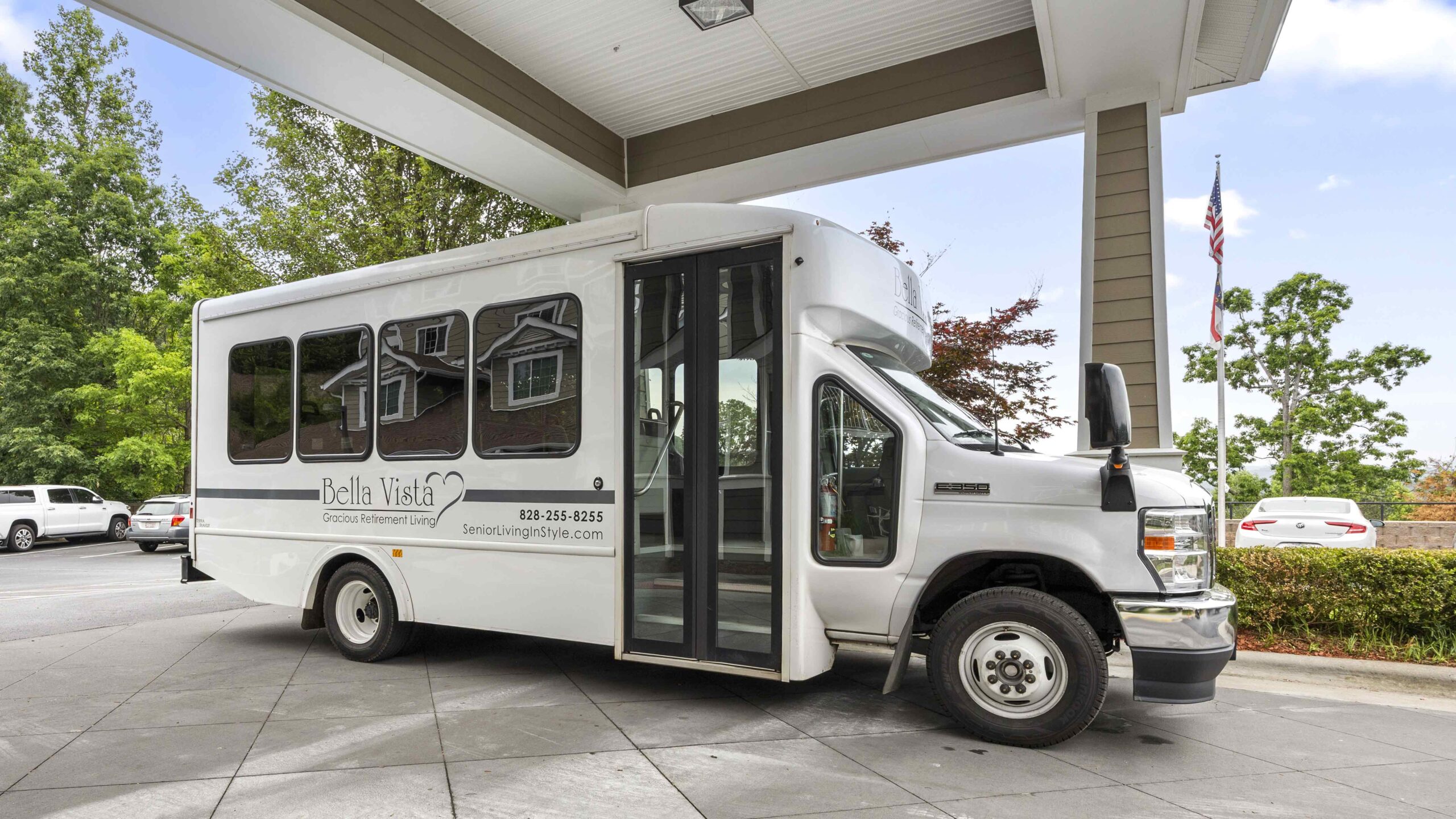 A white shuttle bus labeled "Bella Vista" is parked under a covered driveway near trees and cars, with an American flag in the background.