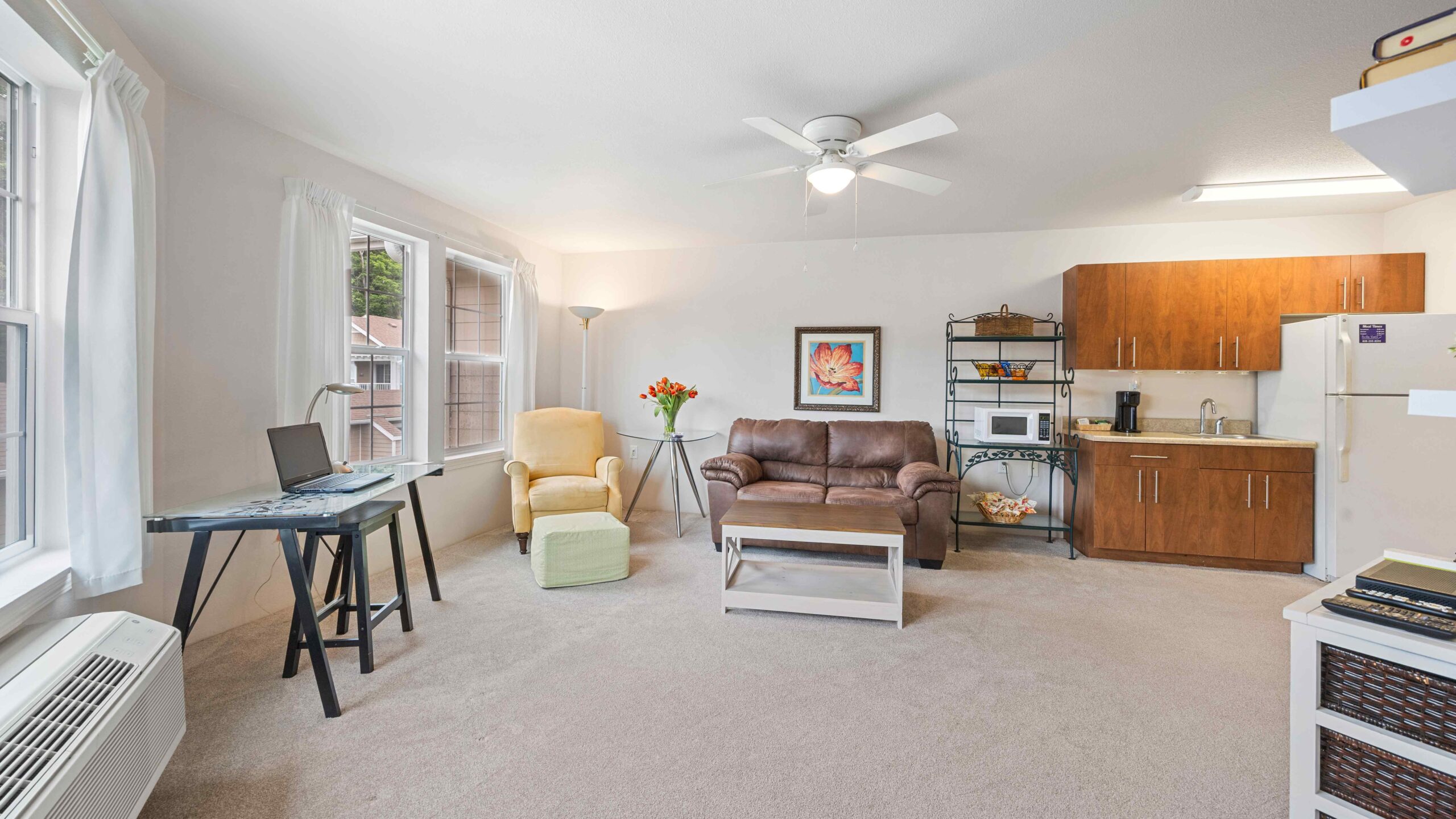 Bright living space with beige carpet, brown sofa, yellow armchair, small desk by the window, coffee table, and kitchen area with wood cabinets, white appliances, and metal shelving. Ceiling fan and wall art are visible.