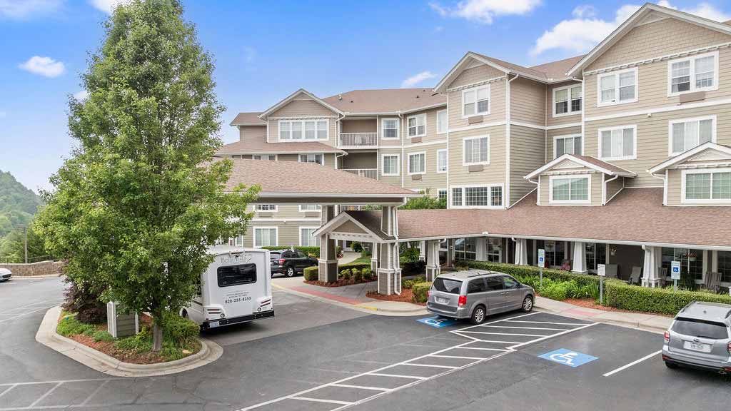 A multi-story beige building with white trim, featuring a covered entrance. Several vehicles, including a van and an RV, are parked in front, with clearly marked handicap parking spaces. Trees and landscaping surround the area.