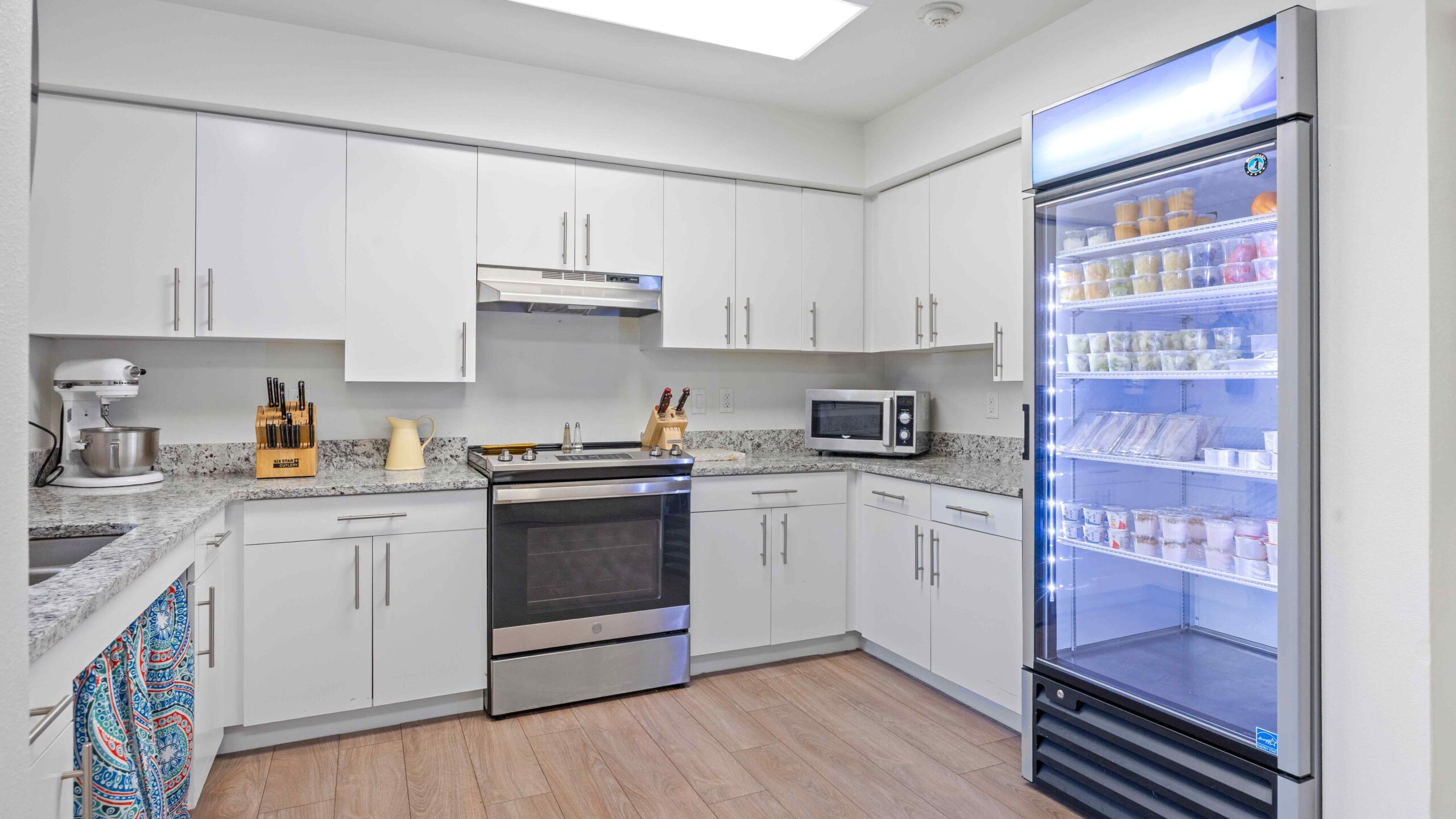 Modern kitchen with white cabinets, stainless steel appliances, a microwave, and a mixer on the counters. A large, lit commercial refrigerator displays drinks and food items on the right side. Hardwood flooring throughout.