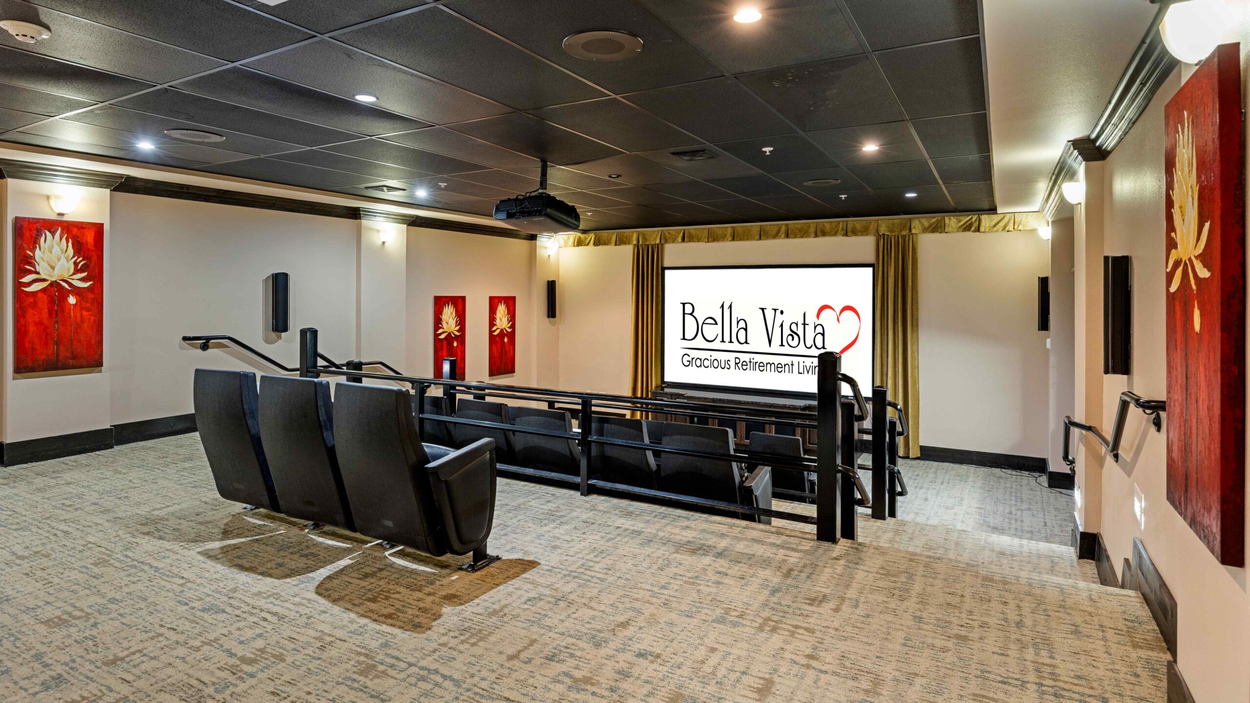 A modern home theater room with black reclining seats facing a large screen displaying "Bella Vista Gracious Retirement Living." The walls are decorated with red floral artwork. The room has soft lighting and beige carpet.