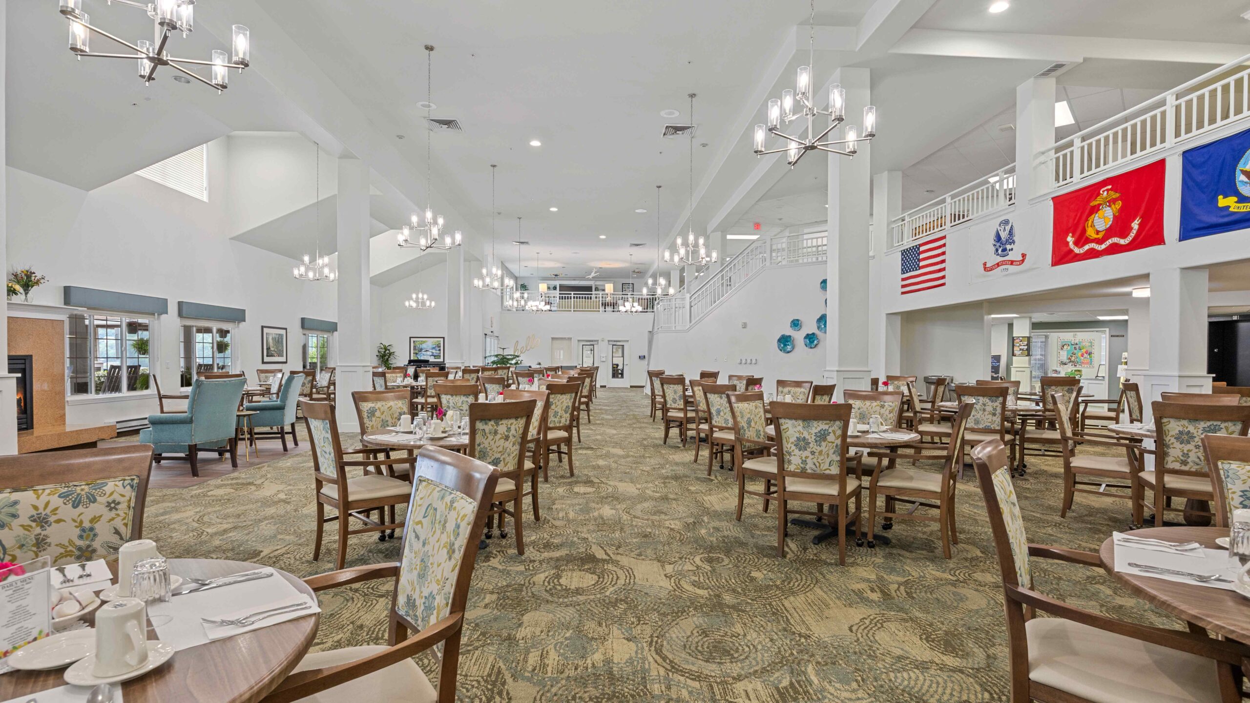Spacious, well-lit dining area with neatly set tables and chairs, chandeliers overhead, and military flags displayed on a balcony railing. Large windows and high ceilings create an open, inviting atmosphere.