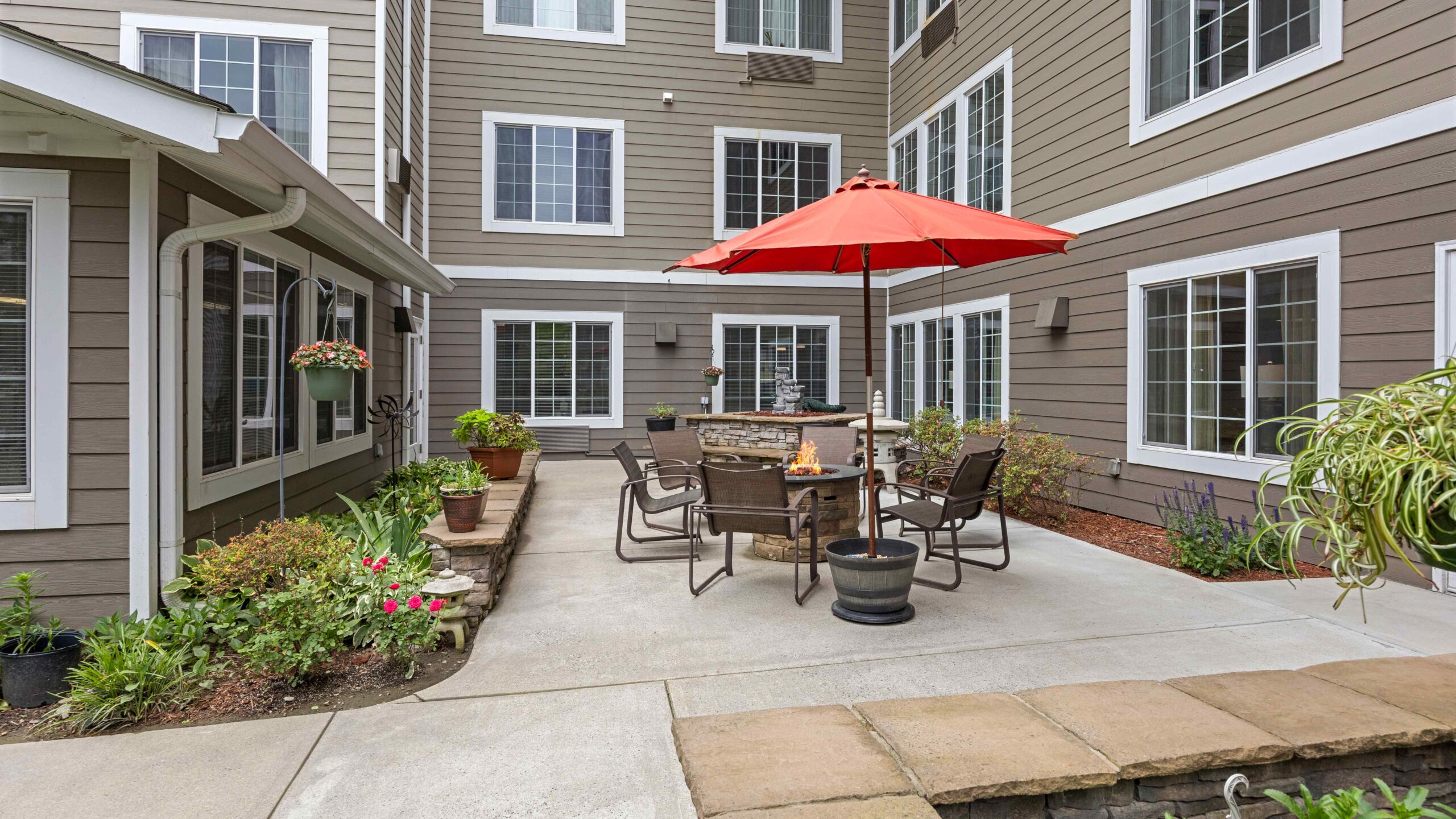 A courtyard with patio furniture, a red umbrella, potted plants, and surrounded by multi-story tan and white buildings with many windows. The area is clean and well-maintained.