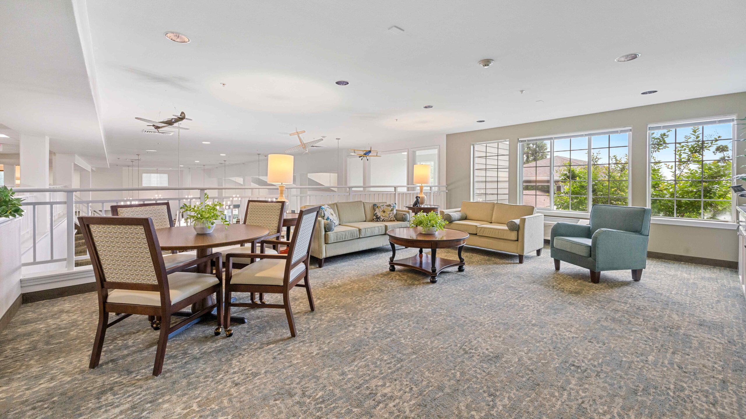 Bright, spacious lounge area with large windows, beige sofas, blue armchair, wooden coffee tables, dining table with four chairs, green plants, and decorative model airplanes hanging from the ceiling.