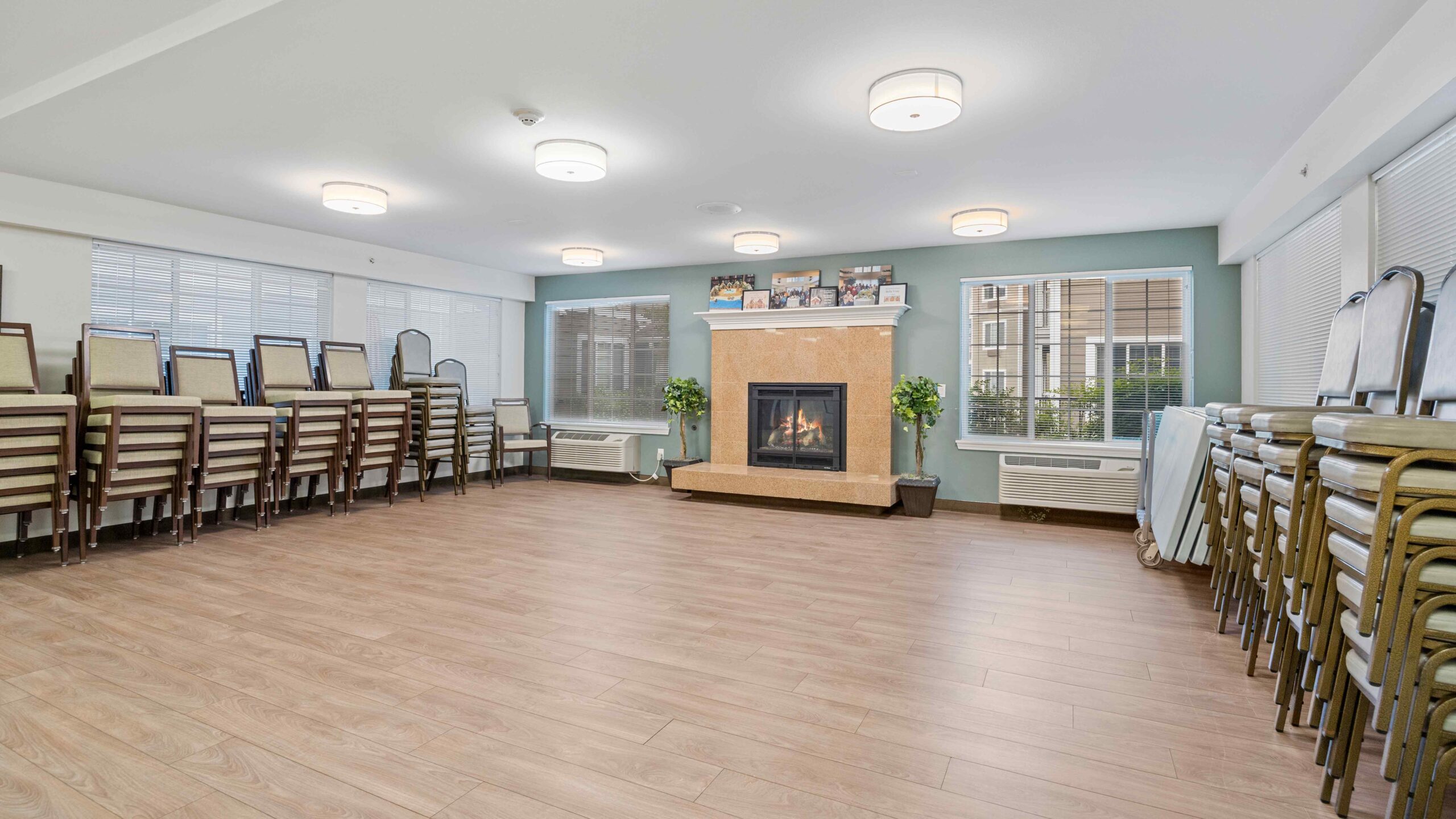 A spacious, well-lit room with a fireplace, light wood flooring, stacked chairs along the walls, and large windows letting in natural light.