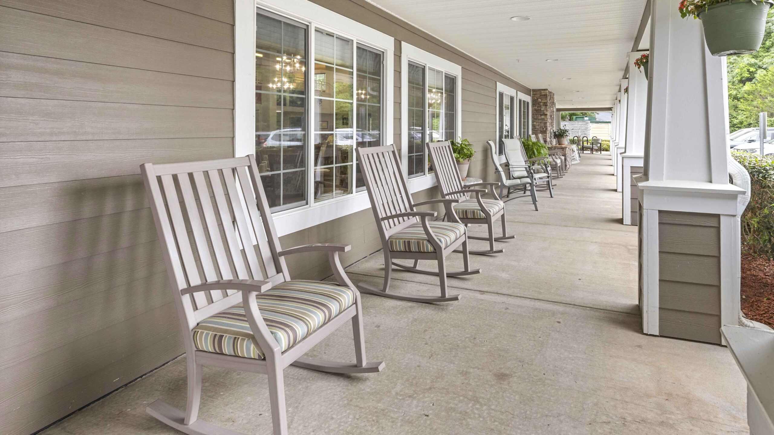 A row of empty rocking chairs with striped cushions lines a covered porch outside a building with large windows. Potted plants hang from the roof and greenery is visible in the background.