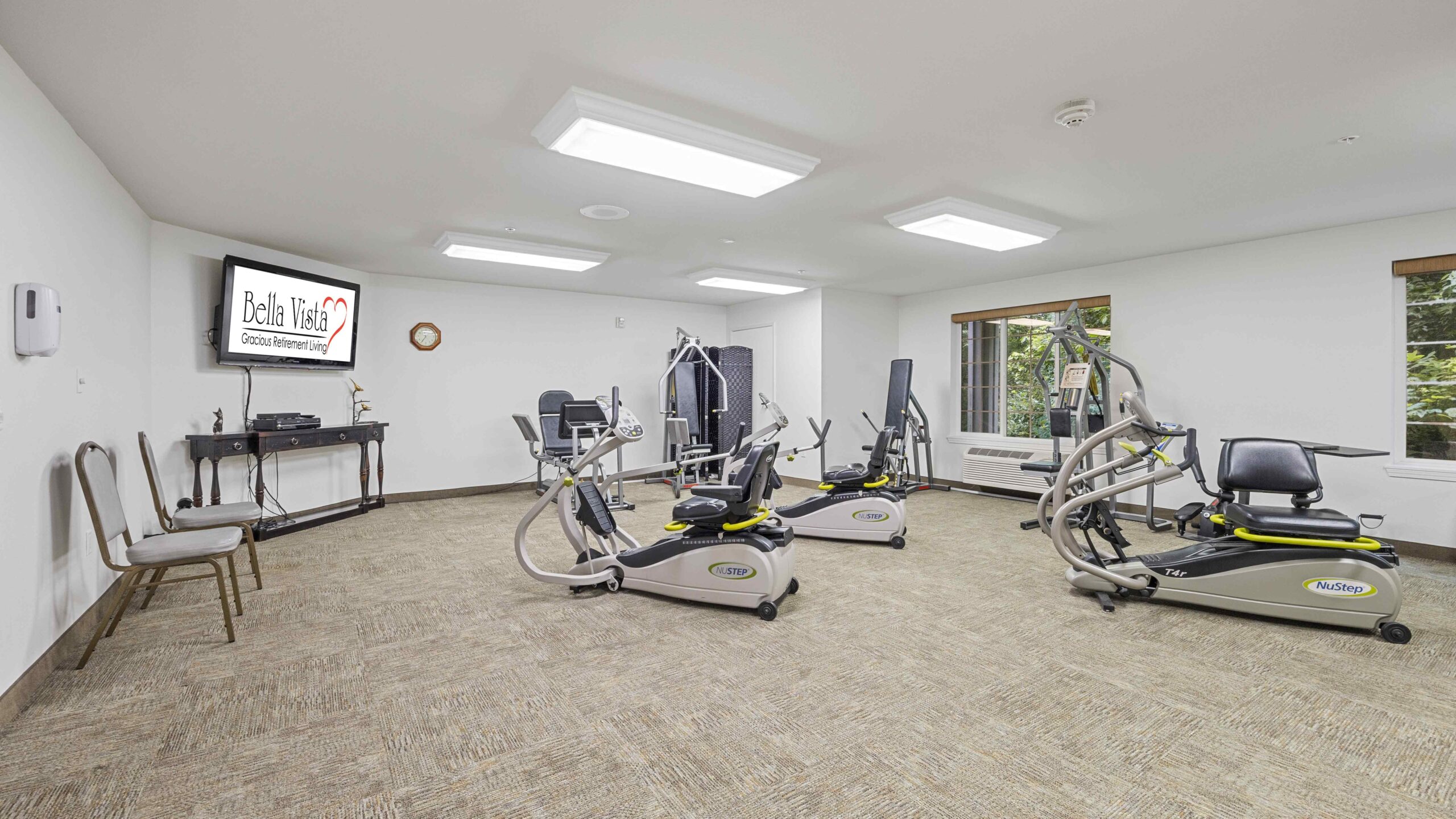 A well-lit exercise room with three pieces of cardio equipment, windows on the right, a TV screen displaying “Bella Vista,” a table with chairs along the left wall, and beige carpeted flooring.
