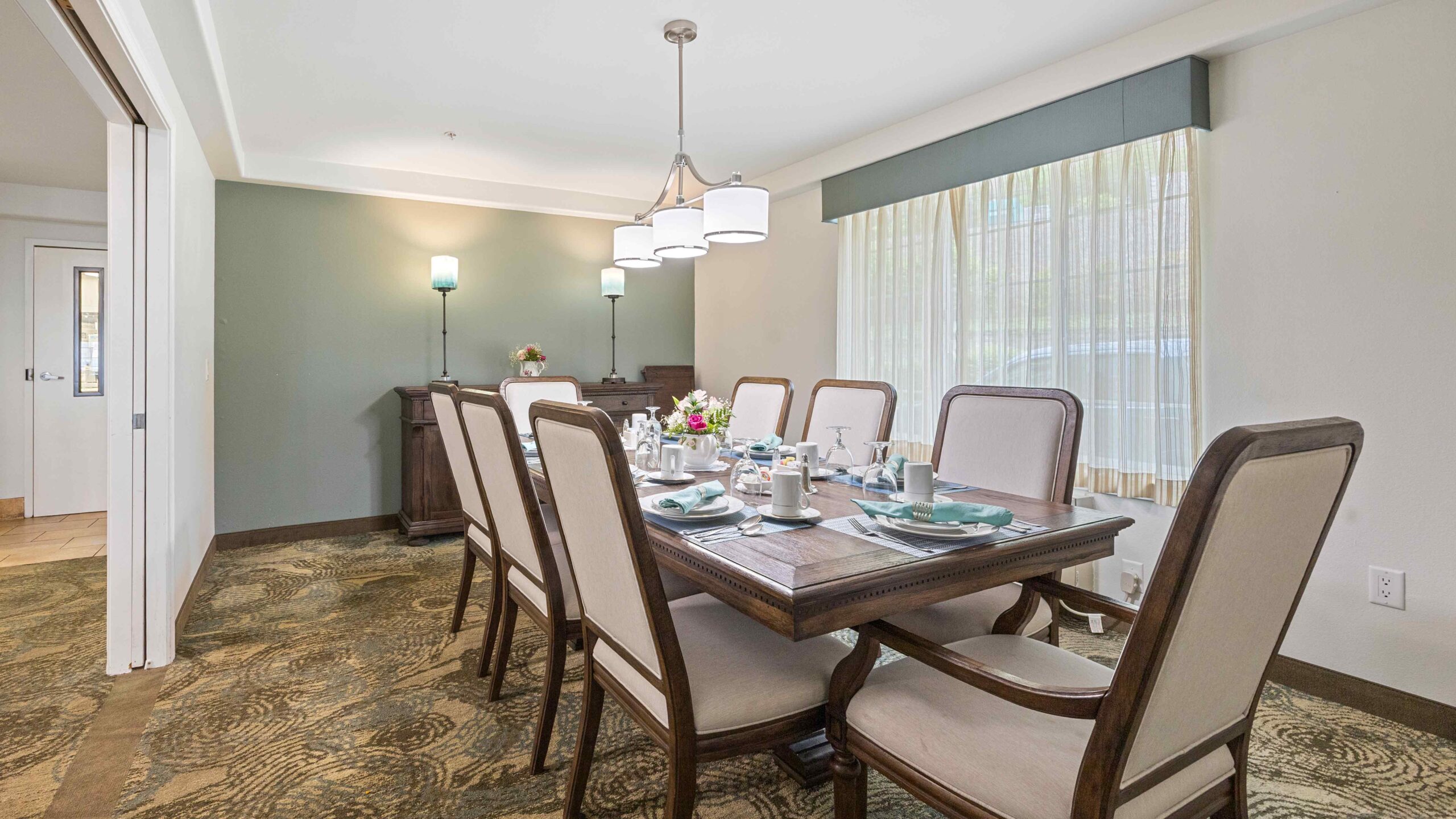 A dining room with a wooden table set for six, cushioned chairs, a sideboard with two lamps, decorative centerpiece, and large window with curtains letting in natural light.