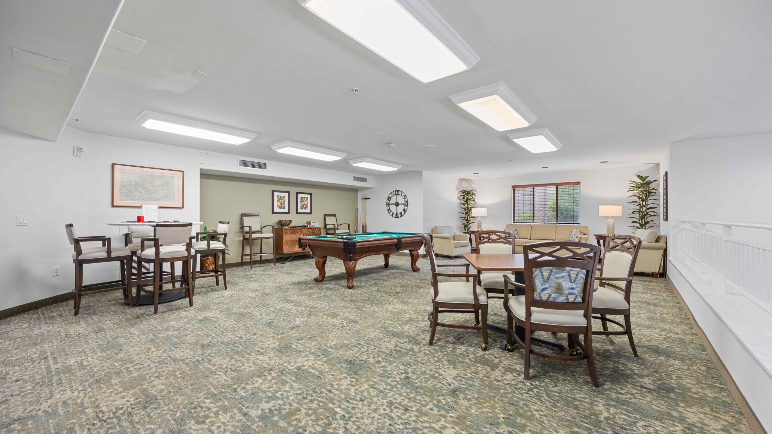 A spacious, brightly lit recreation room with a pool table in the center, surrounded by several tables and chairs, sofas, framed artwork, plants, and large windows letting in natural light.