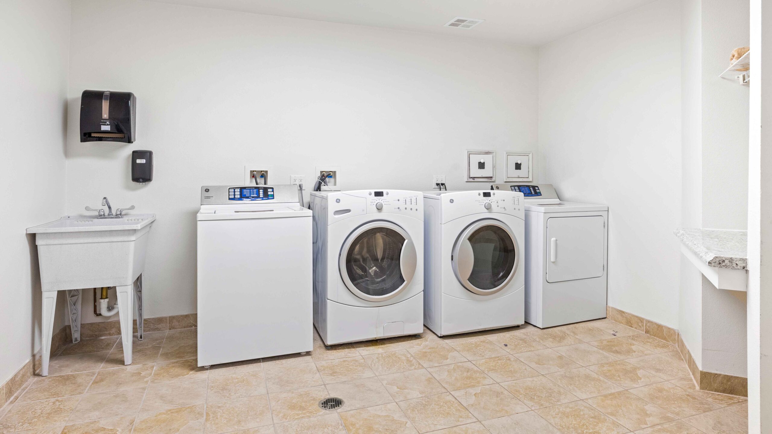 A laundry room with a utility sink, a top-load washing machine, a front-load washing machine, and two front-load dryers, all white, on a tan tile floor with white walls and a wall-mounted paper towel dispenser.