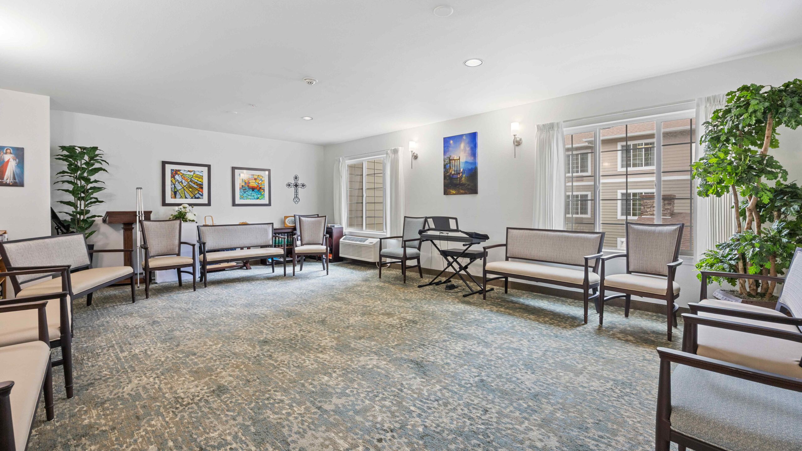 A bright, spacious room with several chairs arranged along the walls, a keyboard, houseplants, and framed artwork on the walls. Large windows let in natural light, and the floor is covered with a patterned carpet.