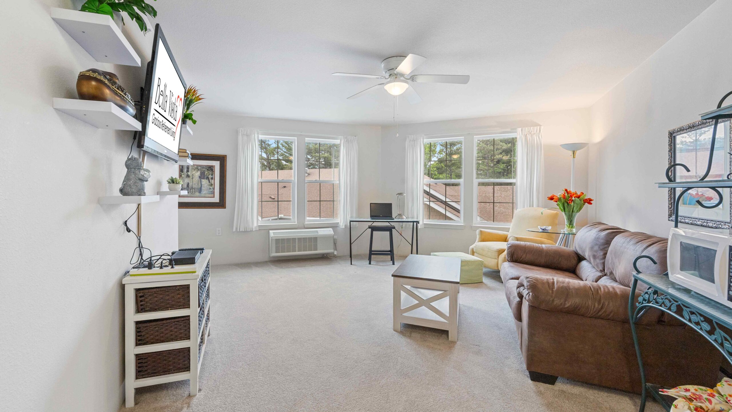 Bright living room with large windows, a brown sofa, a coffee table, wall-mounted TV, small desk with a chair, light-colored carpet, and a shelf with decorative items. A floor lamp and flowers add cozy touches.
